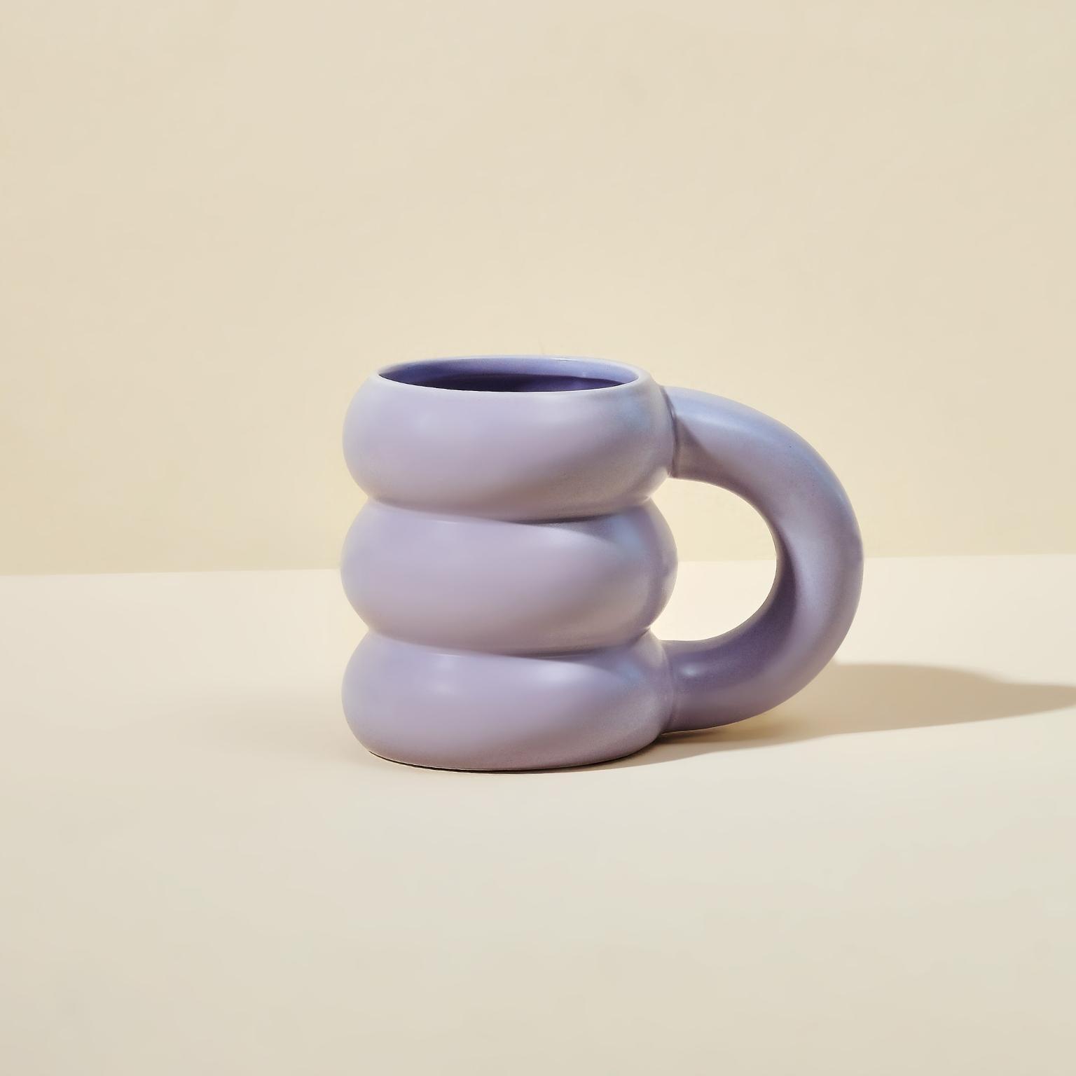 The Blume Cloud Mug is light purple with a sculpted design featuring three rounded sections stacked vertically. It has an oversized handle, offering a modern and artistic look against a plain light beige backdrop.